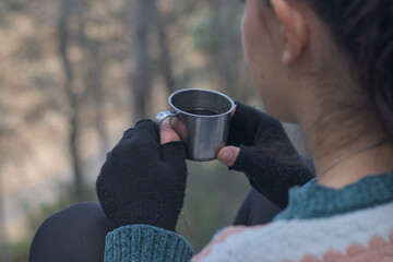 Hand with gloves holding thermal steel mug with coffee in the forest, copy space for text