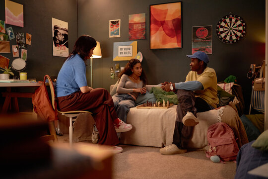 Three friends sitting in cozy dorm room, engaging in chess game near comfortable bed and wall art. Shelves and dartboard adding charming ambiance to room