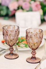 Elegant table setting with brown wineglasses close-up. Fresh pink flowers, cards, dishes, white plates, cutlery on ivory tablecloth
