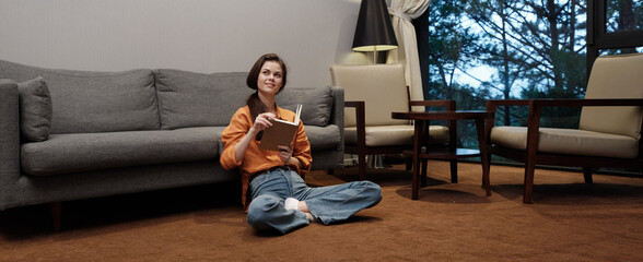 A relaxed young woman reading a book on a cozy carpeted floor in a stylish living room, surrounded...