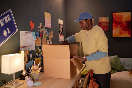 Man packing a cardboard box in his dorm room with various items on desk and wall. Neatly arranged schedule board and motivational poster add personal touch - Powered by Adobe