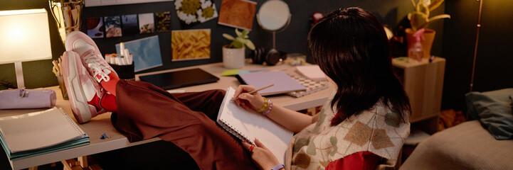Website header shot of person drawing in sketchbook illuminated by table lamp while sitting at desk with plants, sketches, and personal items