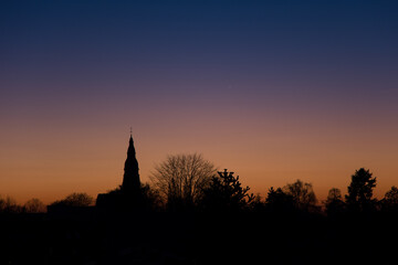 Obraz premium Sunset at the golden hour with the silhouette of the Christuskirche and a leafless tree in Ratingen Homberg with orange and blue sky