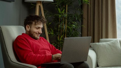 A young man in a red sweater sitting on a couch, reacting with excitement and surprise while using a laptop