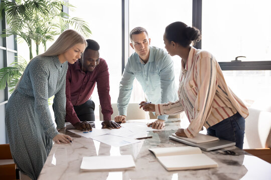 Young diverse teammates engaged in brainstorming, decision-making during meeting in modern boardroom, reviewing infographic data laid out on conference table, collaborate on strategy or joint project