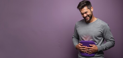 Man holds heating pad on stomach, experiences abdominal pain. Facial expression shows suffering, discomfort. Medical concept for stomachache, intestinal spasms or colic. Purple backdrop has copyspace.