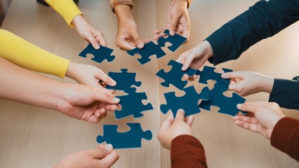 Diverse hands reach out to assemble blue puzzle pieces on a wooden table, symbolizing teamwork and...