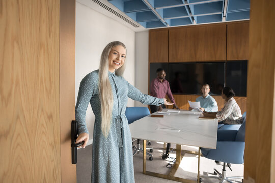 Smiling businesswoman stands at door of modern conference room, making inviting gesture and welcoming partner or client to join meeting, while group of colleagues engaged in discussion in background