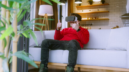 Young man wearing VR headset, fully immersed in virtual reality experience, gesturing with hands while sitting on cozy couch in modern living room