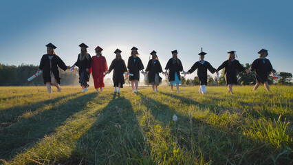 Group of cheerful graduates walking together in a field at sunset, holding hands and diplomas,...