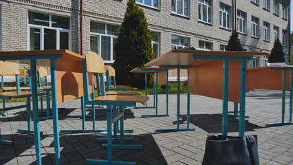 Empty school desks and chairs are arranged outdoors in front of a school building, suggesting an outdoor class or break time