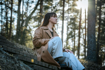 A woman in a trench coat and light jeans sits thoughtfully on forest steps in soft sunlight, surrounded by tall trees with a subtle lens flare.