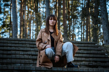 Woman in a tan trench coat and light jeans poses assertively on outdoor stone steps, surrounded by forest trees and golden-hour sunlight.
