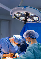 Surgeons operating in hospital. Two surgeons in scrubs operate under bright lights while focused on...