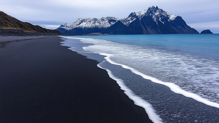 Dark sand beach meets a vibrant sea, snow-capped mountains loom