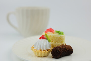Close up side view of mini desserts lying on a plate, behind them is a white mug with a hot drink on a white background with space for text. The concept of mini desserts, sweets, confectionery. High