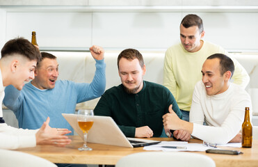 Group of male friends gathering at home enthusiastically celebrating good news, seen on laptop screen, with lively gestures and drinks..
