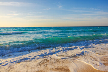 Waves rolling onto sandy beach with emerald Caribbean sea under clear blue sky in golden evening light. Mexico.