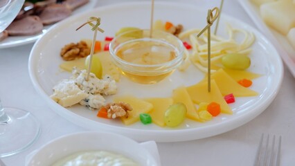 Gourmet cheese selection featuring multiple varieties, accompanied by honey, grapes, walnuts, candied fruits on pristine white serving plate