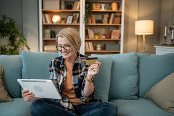 Smiling senior woman using digital tablet and credit card for online shopping on sofa at home