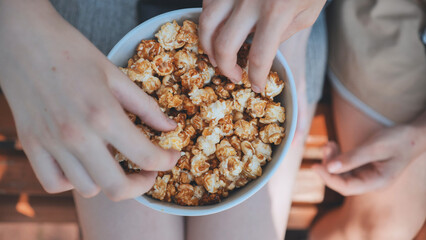Hands diving into caramel popcorn while relaxing on wooden park bench during bright summer afternoon