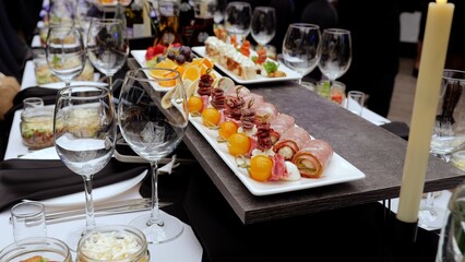 Elegant table setting featuring stemmed wine glasses, gourmet canapes, meat rolls, and fruit skewers near glowing candlelight, showcasing upscale event preparation
