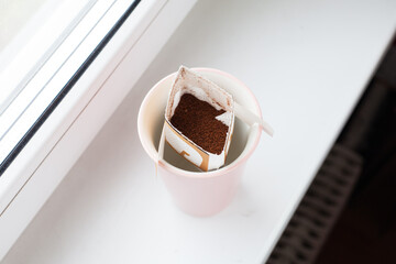 Drip bag in a ceramic cup on windowsill. Instant freshly brewed alternative filter drink in mug. Easy brew black coffee method for home, travel or office. cozy trendy lifestyle 