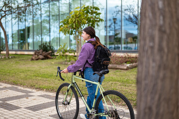 Young man walking bicycle next to modern building with laptop backpack.