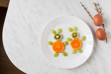 Creative healthy snack of breakfast for kids fruit bunny shaped on white plate, easter eggs around. Rabbit, basket makes from grape, orange, banana, apple. Top view