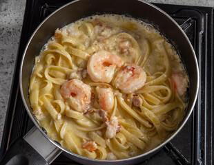 Top view of a pot of shrimp with bacon bits and fettuccine or linguini on a black stove