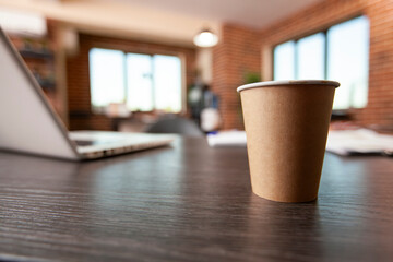 Selective focus on brown disposable mug placed on office desk with laptop nearby. Closeup of paper cup arranged on clean table with digital device in vacant brick wall office.