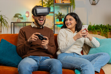 Happy Indian man wearing VR goggles playing videogame with joystick sitting beside Indian...