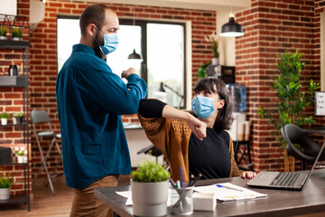 In bright office, businessman and woman exchange elbow bumps as part of safe communication routine. Caucasian coworkers planning and discussing work at desk, ensuring pandemic safety guidelines.