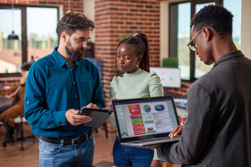 Businessman and woman reviewing online documents on tablet as black man checking business analytics on his laptop. Male and female coworkers using digital devices in brick wall office.