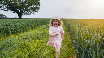 Happy child sticking out tongue while running through green field near big oak tree in summertime