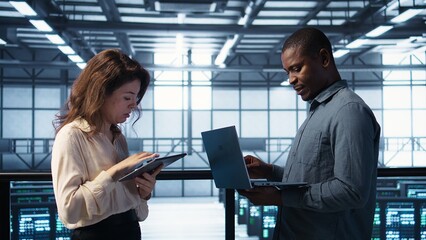 Developers in data center using laptop and tablet, executing security patches, securing firewall. Server farm workers using devices to perform vulnerability scans, ensuring data protection, camera B