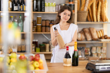 Polite young girl seller is kindly offering a bottle of wine in grocery store
