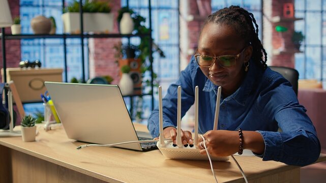 African american woman restarting her wireless router and checking network settings to resolve an internet error disrupting her daily tasks. Frustration due to signal loss. Camera A.