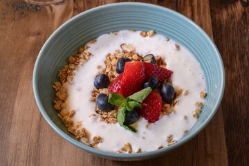 Blue bowl of breakfast granola with yogurt, fresh strawberries and blueberries on rustic wooden table