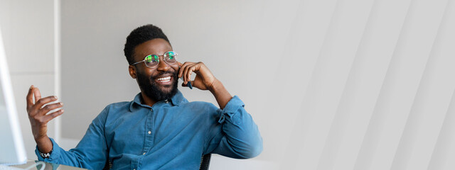 A young African American entrepreneur smiles while talking on the phone in a modern office. He...