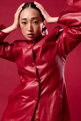 Fashion asian woman in red leather jacket posing with hands on head against red background