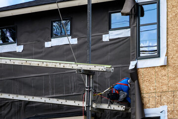 New housing development home construction, workman on scaffolding installing moisture resistant membrane on home, sunny winter day on job site
