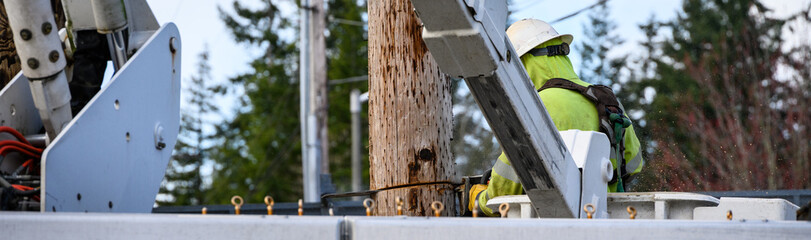 Closeup of power utility lineman in lift bucket with chainsaw cutting off temporary wood pole, professional at work
