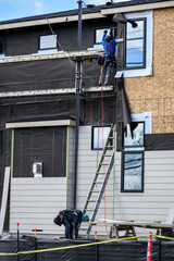 New housing development home construction, workman on scaffolding installing moisture resistant membrane on home, sunny winter day on job site
