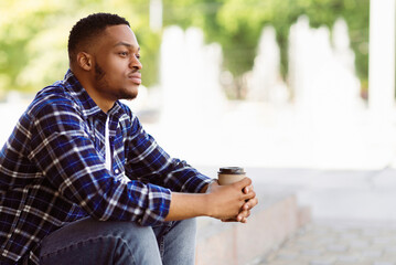 Fototapeta premium Side view of pensive african american person sitting in the park with coffee, looking aside, free space