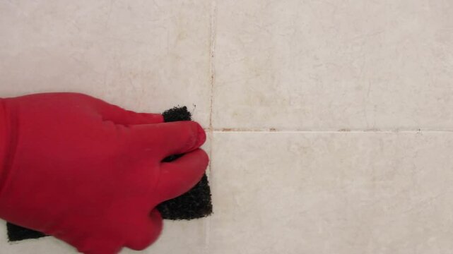 Mano con guante de latex rojo tallando azulejo de ba&ntilde;o con fibra. Limpieza de ducha y regadera.