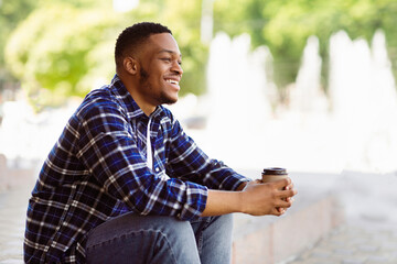 Fototapeta premium Enjoying Free Time. Cheerful black guy sitting on the stairs, holding cup of coffee, copy space