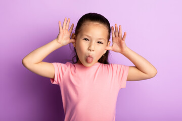 Portrait of cute asian kid sticking out her tongue and goofing at studio, isolated on pastel purple...
