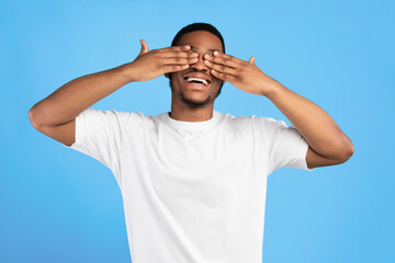 Naklejka premium Cheerful African American Guy Covering Eyes With Hands Standing On Blue Background In Studio. I Don't Look Concept