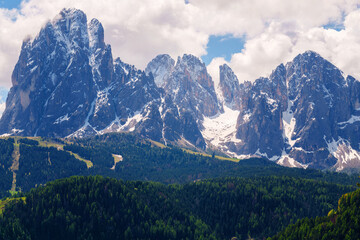 Majestic mountain range with snow-capped peaks and lush forests under a cloudy sky near Valley of Funes at Dolomites, Italy.
